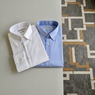 Das Bild zeigt einen Tisch. Darauf liegen nebeneinander ein weisses und blaues Button Down Hemd für Herren