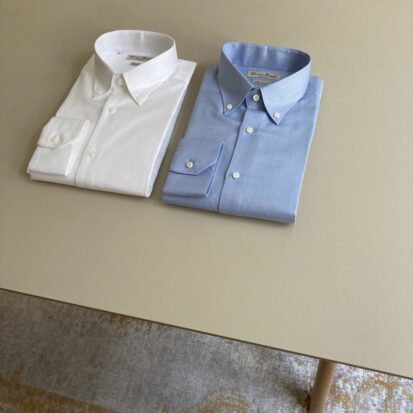 Das Bild zeigt einen Tisch. Darauf liegen nebeneinander ein weisses und blaues Button Down Hemd für Herren